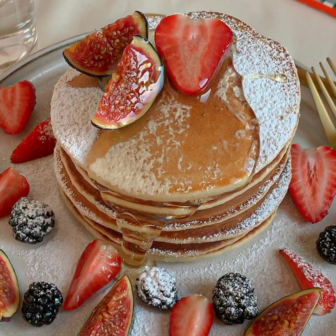 A stack of fluffy pancakes topped with fresh strawberries, figs, powdered sugar, and a drizzle of syrup, surrounded by blackberries, strawberries, and figs on a plate.